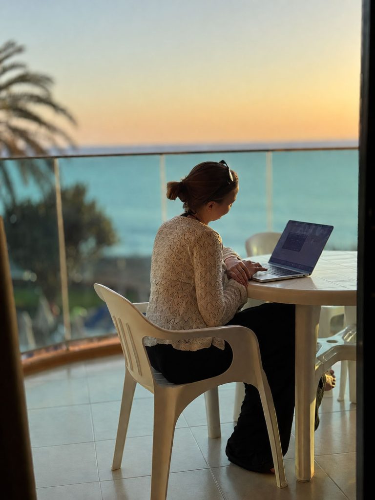 Person som arbeider på laptop utendørs med utsikt over havet ved solnedgang.